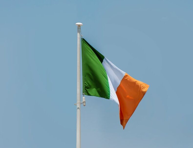 Ireland national flag on a flagpole waving against a clear blue sky.